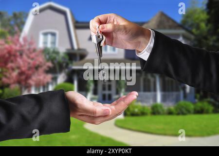 A hand giving a key to another hand. Both persons in suits and a house in the background Stock Photo