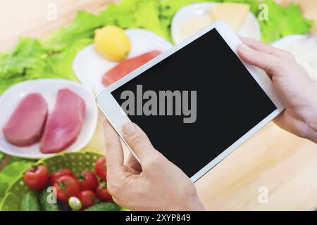 Close-up top view of female hands holding a tablet computer with a blank display surrounded by healthy food on a home kitchen table. Workpiece to inse Stock Photo