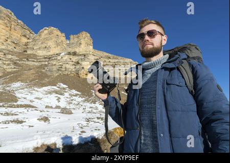 Portrait A bearded hipster photographer with a backpack and wearing sunglasses with a large backpack on his shoulders stands with a DSLR camera in his Stock Photo