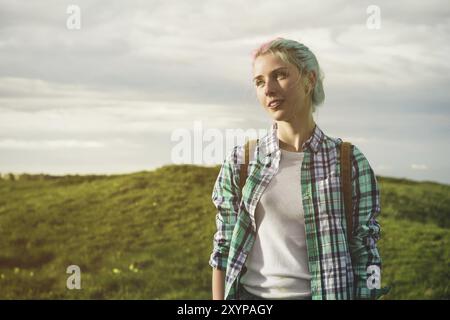 Portrait of a young smiling attractive woman in plaid shirt clothes with small backpack at sunny day on the cloudy sky and field background. joyful wo Stock Photo