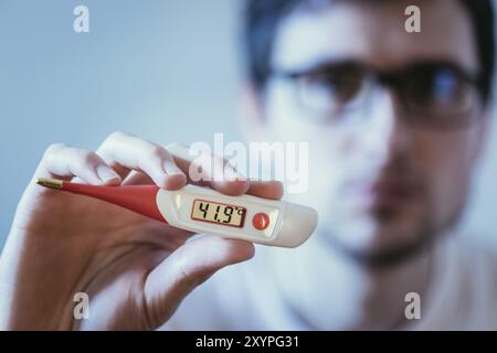 Man holds a red fever thermometer with high temperature in his hand, blurry face Stock Photo