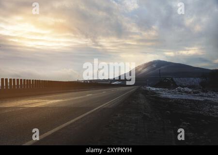 A country asphalt road illuminated by the rays of the sun through the clouds leading to the mountain on which the rays of the sun fall in the evening Stock Photo
