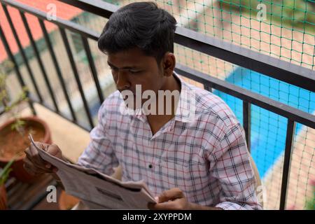 A young man, dressed in a checkered shirt, sits on a balcony reading a newspaper. Potted plants surround him as he enjoys the peaceful atmosphere and Stock Photo