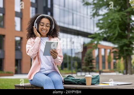 Smiling African American female student wearing headphones using tablet outside modern building. She seems relaxed and engaged, sitting on bench, enjoying music or studying. Stock Photo