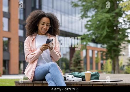 Smiling African American female student using smartphone while sitting outdoors. Relaxing during study break with coffee and headphones nearby, conveying a sense of happiness and relaxation Stock Photo