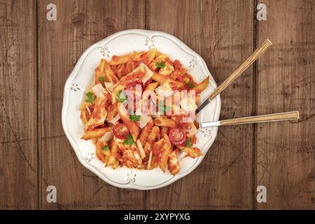 An overhead photo of a plate of penne pasta with chicken and tomato sauce, with a fork and a spoon plunged into the dish, shot from above on a dark ru Stock Photo