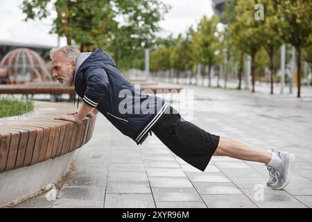 Side view of fit elderly man in sportswear doing push ups on bench working out regularly in city park with tiles, copy space Stock Photo