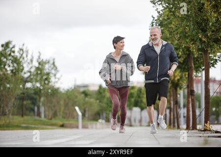 Wide angle shot of fit elderly woman and man in sportswear smiling while jogging together outside along lane of trees working out regularly in morning park, copy space Stock Photo