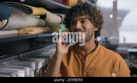 Arabian Indian man happy customer salesman fashion stylist talking mobile phone shopping in fabric store cheerful male look at fiber linen silk satin Stock Photo