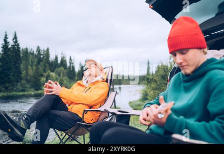 Peaceful Norwegian camping scene with father and son by lakeside. Father relaxes in chair while son plays ukulele surrounded by nature, they enjoy qui Stock Photo