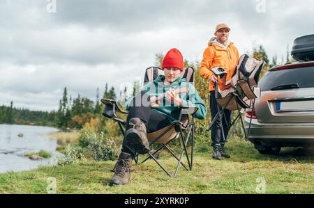 Peaceful Norwegian camping scene with father and son by lakeside. Teen boy playing ukulele surrounded by nature enjoying moment together in their car Stock Photo