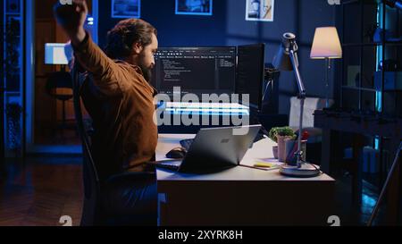 Fatigued developer working overnight at home, rubbing eyes while taking break from coding, feeling sleepy. Tired IT admin exhausted from too much screen time, pausing tasks to rest, camera B Stock Photo