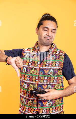 Portrait of annoyed man showing thumbs down sign holding gamepad, disapproving of videogames. Disgusted person protesting against gaming, doing rejection hand gesture, isolated over studio backdrop Stock Photo