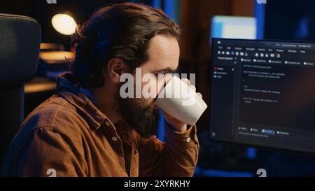 Focused programmer drinking coffee in apartment while solving database errors on computer. Remotely working IT admin concentrated on finalizing coding project in home office, camera A Stock Photo