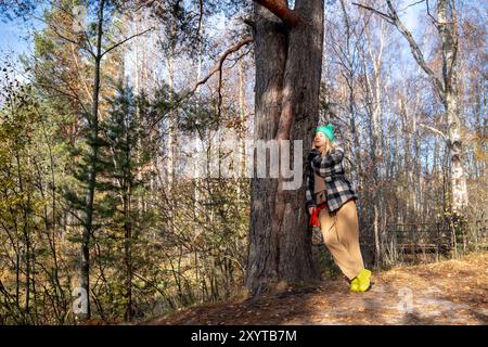 Carefree woman rest after hiking, leans to tree, thinks dreams in autumn forest. Nature therapy Stock Photo