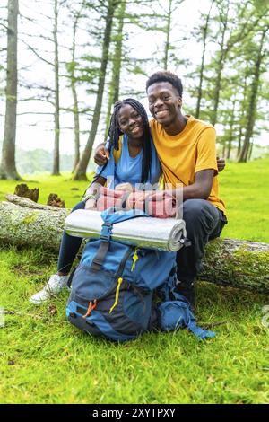 Vertical portrait of a cute young african couple embracing sitting on a log in a green beauty forest in spring Stock Photo