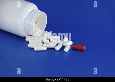 Overturned white medicine bottle with its contents white pills or capsules, spilled out onto a blue background. Among these white pills, a solitary re Stock Photo