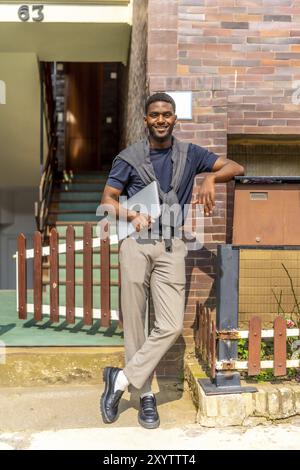 Vertical full length portrait of an african young businessman carrying a laptop outdoors next to a residential building Stock Photo