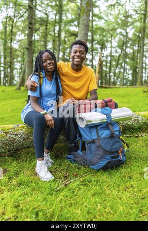 Vertical portrait of young african friends embracing sitting on a log together in a green forest in spring Stock Photo