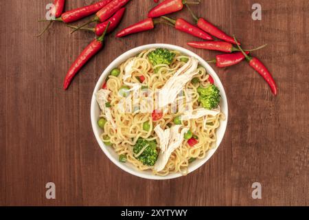 An overhead photo of a bowl of noodles with chicken, green peas, and broccoli, with red hot chili peppers and copy space, on a dark rustic background Stock Photo