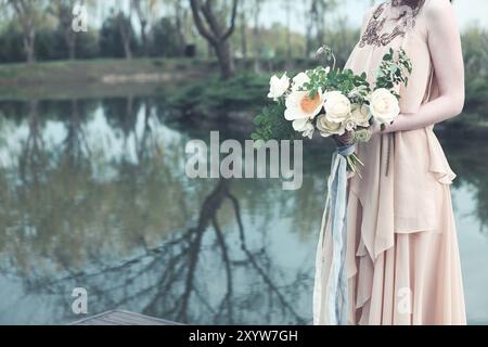 Crop woman wearing boho chic pink long dress and holding gorgeous bouquet with ribbons on pier in park Stock Photo