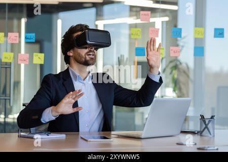 Businessman engaged with virtual reality headset in an office setting. Immersive experience demonstrates technological innovation. Professional appears intrigued and focused, surrounded by office Stock Photo