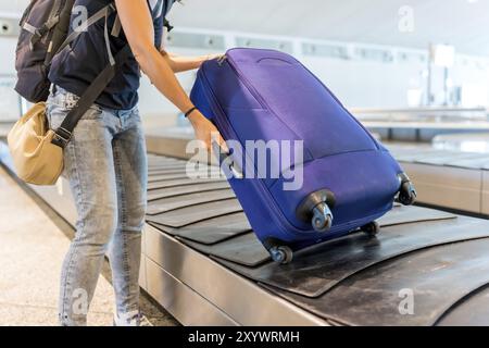 A woman is pulling her blue suitcase off the conveyor belt at an airport. She is wearing a blue shirt and jeans Stock Photo
