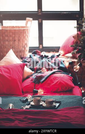 Tray with cups of coffee served on bed with red and green blanket and pillows in Christmas morning in cozy bedroom with garland Stock Photo