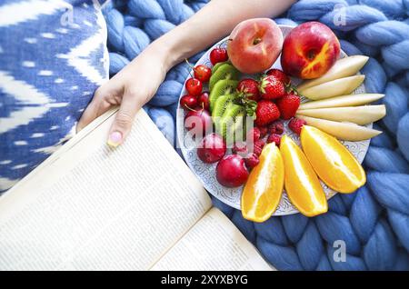 Plate with fresh fruit salad on a blue plaid and book. Woman reading the book and eating fresh fruits. Close up. Top veiw. Cozy and healthy concept Stock Photo