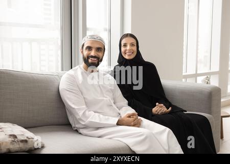 Cheerful Muslim couple in Arabic clothes sitting on home sofa Stock Photo