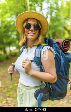 Vertical portrait of a young woman wearing a backpack smiling while walking through a forest path Stock Photo