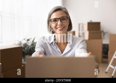 Smiling, professional middle-aged woman extending cardboard box towards viewer Stock Photo