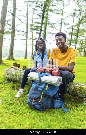 Vertical portrait of two young male and female african friends smiling while sitting on a log in a beauty forest Stock Photo