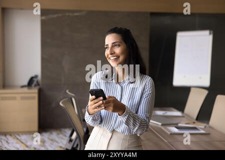Cheerful dreamy Latin executive woman using mobile phone, digital gadget Stock Photo
