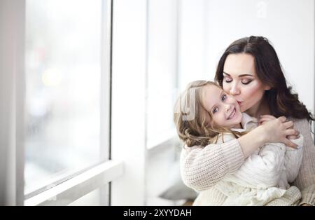 Happy loving family together. Mother and child daughter hugging by the window wearing cozy sweaters Stock Photo