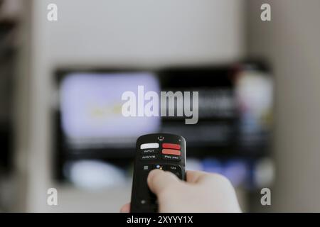 Hand holding remote control pointing to TV. Person watching smart television indoors. Blurred background. Colorful screen, copy space. Person browsing Stock Photo