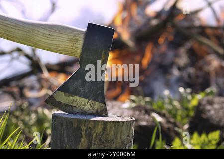 Axe embedded in the stump of a tree in close up, burning campfire in the background, outdoor activities such as chopping wood, splitting logs or envir Stock Photo