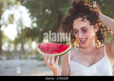 Beauty smiling curly woman is wearing pink sunglasses and eating watermelon on the beach Stock Photo