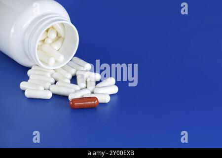 Overturned white medicine bottle with its contents white pills or capsules, spilled out onto a blue background. Among these white pills, a solitary re Stock Photo