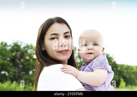 Cheerful female holding adorable baby up standing against lush green fields under blue sky Stock Photo