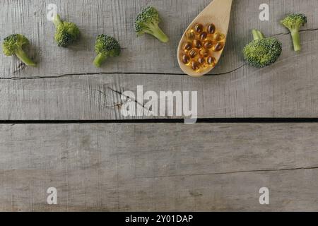 Flat lay with omega 3 capsules on wooden spoon, broccoli on wooden background. Natural medicine, health concept. Vitamin pills. Stock Photo