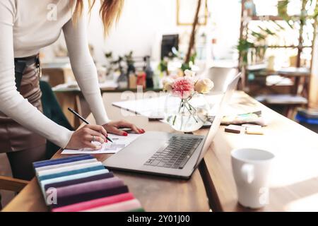 An interior designer is working on a new project. Creating a sketch. Choosing color from fabric color palette. Oak wooden table. Cup of coffee, Flower Stock Photo