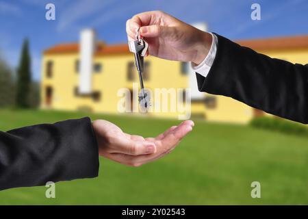 A hand giving a key to another hand. Both persons in suits. House in the background Stock Photo