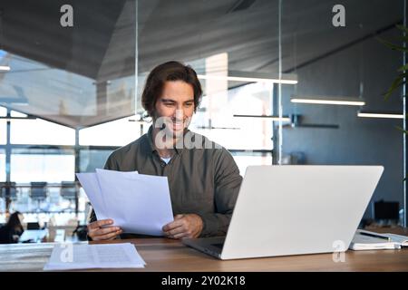 Happy busy business man reading documents looking at laptop working in office. Stock Photo