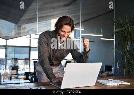 Excited young business man winner celebrating financial success using laptop. Stock Photo