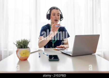 Young beautiful female doctor wearing headphones, making online webcam video call on laptop screenand gives advice to patient. Videocall view. Tele me Stock Photo