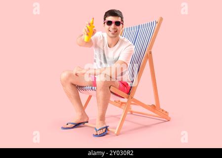 Young man with sunscreen cream sitting in deck chair on pink background Stock Photo