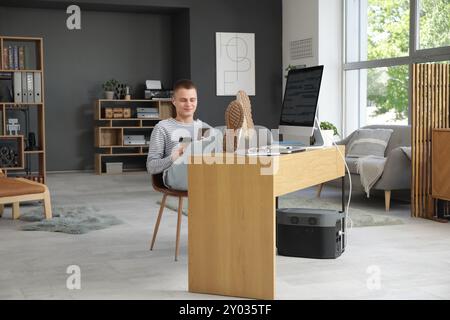Young male programmer with mobile phone and cup of coffee relaxing at his workplace in modern home office Stock Photo