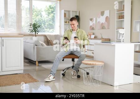 Young male freelancer with mobile phone and cup of coffee relaxing at his workplace in light home office Stock Photo