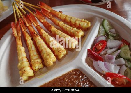 A close-up shot of delicious grilled satay shrimp skewers served with a side of peanut sauce and a fresh cucumber salad garnished. Stock Photo